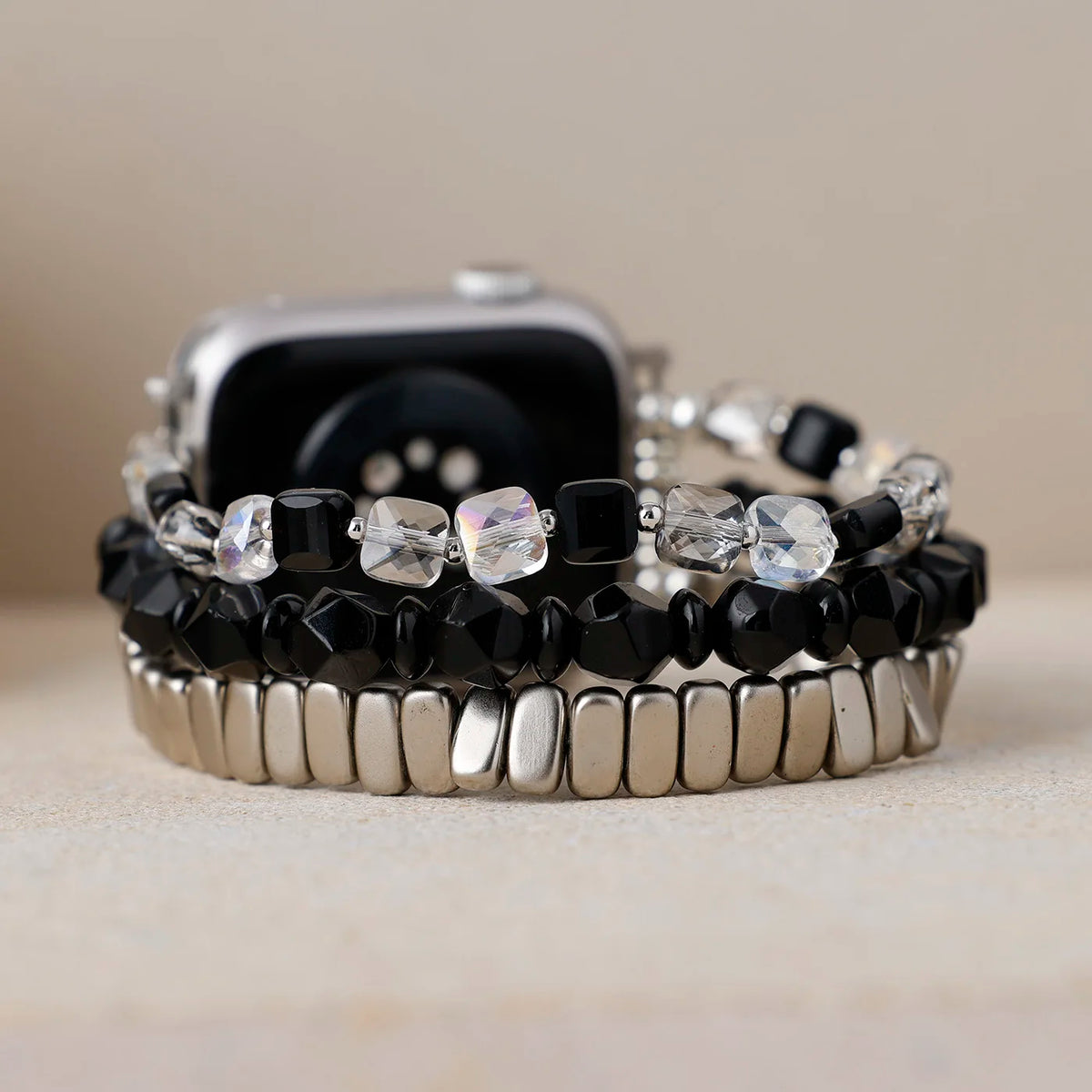 A stack of three beaded watch bands displayed on a silver smartwatch, featuring black faceted beads, clear rhinestone accents, and polished silver-toned metal links, set against a neutral beige background.