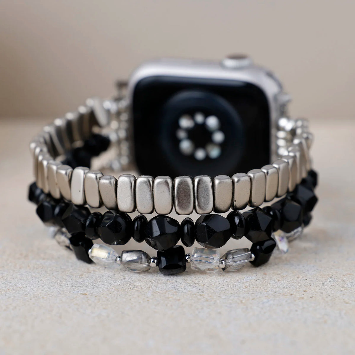 A close-up of a silver metal watch band with three coiled layers: a top layer of polished silver rectangular beads, a middle layer of faceted black beads, and a bottom layer of alternating clear and silver faceted beads. A silver smartwatch is partially visible behind the band.