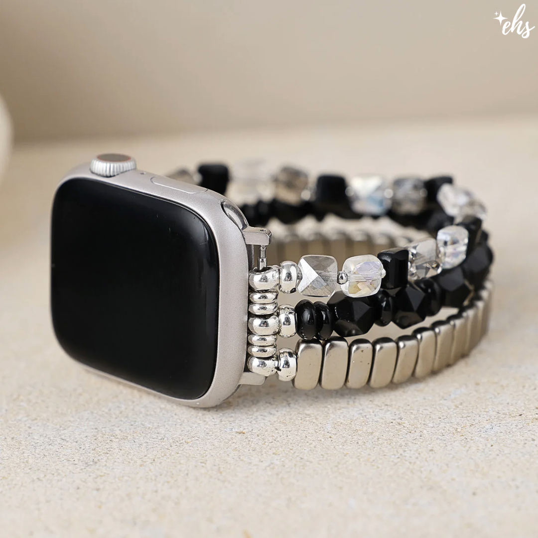 A silver smartwatch with a black screen and a decorative beaded band featuring black faceted beads, clear rhinestones, and metallic silver links, displayed on a light beige textured surface.