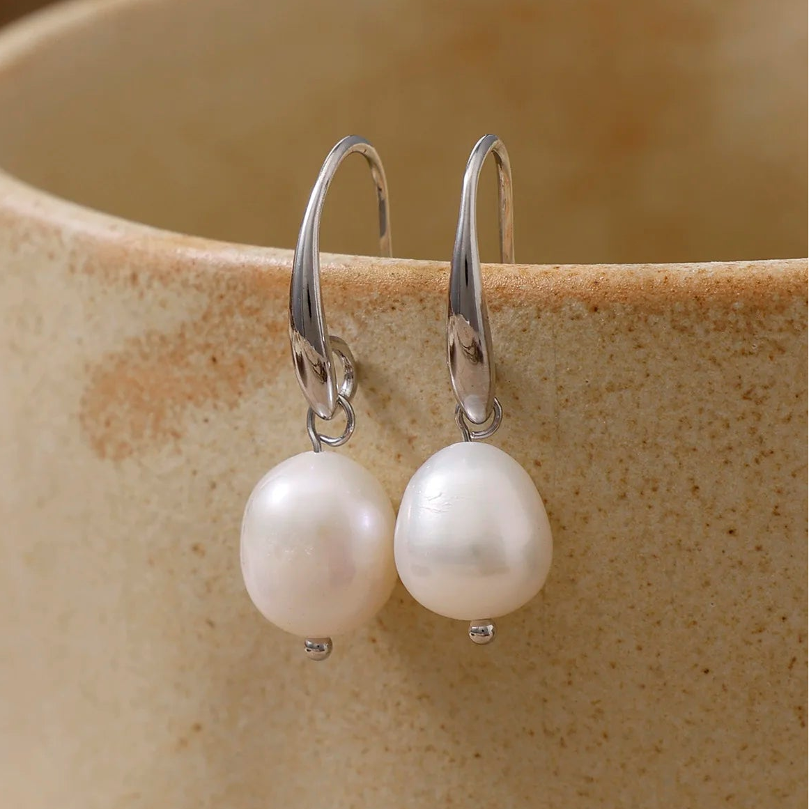 A pair of silver drop earrings with irregular white freshwater pearls, hanging from the rim of a speckled beige ceramic mug.