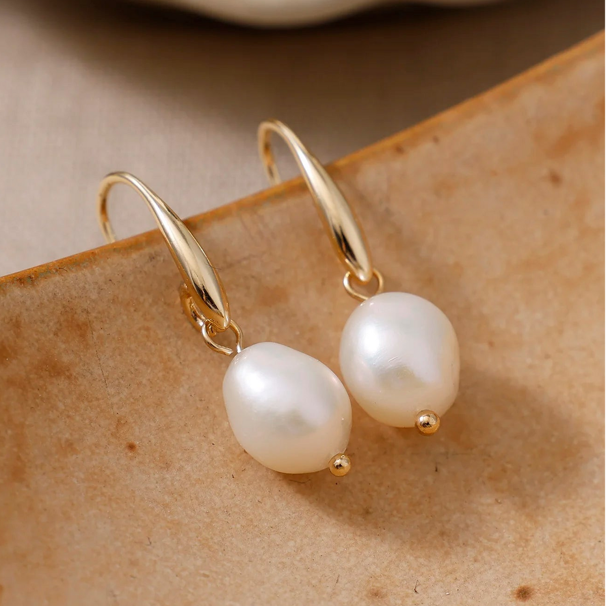 A pair of gold drop earrings with irregular white freshwater pearls, resting on a textured brown surface.