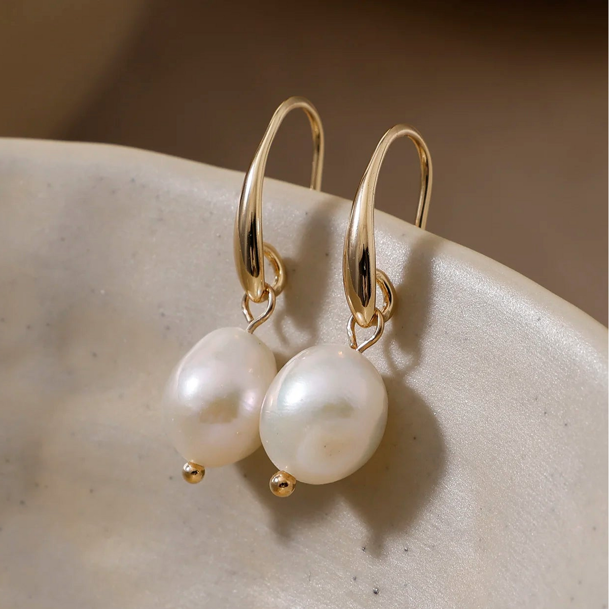 A pair of gold hoop earrings with irregular white freshwater pearls, resting on a textured beige ceramic dish.
