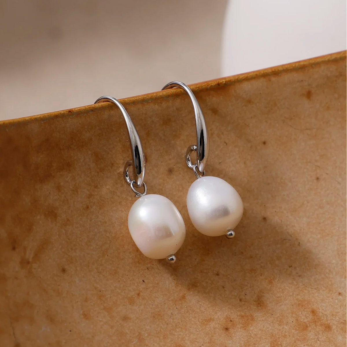 A pair of silver huggie hoop earrings with irregular white freshwater pearls, displayed on a textured brown ceramic dish.